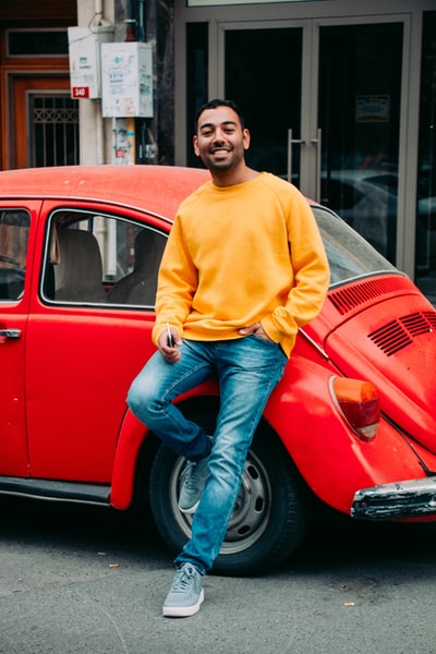 Wearing a yellow sweater and a blue denim jeans man sat on a red Volkswagen beetle
